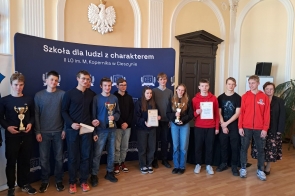 Grupa młodych osób i dwie dorosłe kobiety pozują w eleganckiej sali z jasnymi ścianami i godłem Polski. Część osób trzyma puchary i dyplomy. W tle granatowy baner z napisem o szkole. Wszyscy uśmiechnięci, atmosfera oficjalna, wyraźnie celebrująca sukces.
