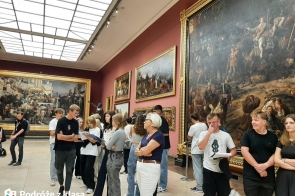 Grupa dzieci zwiedzających galerię sztuki, ogląda monumentalne obrazy o tematyce historycznej. Dyskutują między sobą lub z nauczycielką, która im towarzyszy. Kilka osób trzyma materiały edukacyjne, patrząc się na obraz. Ściany galerii zdobią duże obrazy, w złotych ramach, przedstawiające sceny bitewne i historyczne. Jasne, przeszklone sufity odprowadzają naturalne światło.
