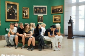 Grupa młodych ludzi siedzi na ławce w galerii sztuki w Krakowie, otoczona obrazami o treści historycznej, zawieszonymi na zielonych ścianach. Pomieszczenie jest przestronne, dobrze oświetlone, dzięki dużym oknom. W dolnym rogu znajduje się logo „Podróże z klasą”, co oznacza, że uczestnicy wycieczki są uczestnikami programu „Podróże z Klasą”.