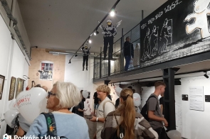 Na zdjęciu widoczni są uczniowie zwiedzający muzeum, w którym znajdują się prace artystyczne Banksy'ego. Na ścianach znajdują się charakterystyczne dla Banksy'ego murale, w tym postacie aniołów oraz inne elementy street artu. Grupa ludzi ogląda ekspozycję, w rękach trzymają balony z logo "Muzeum Banksy'ego". W tle widać także balustradę.