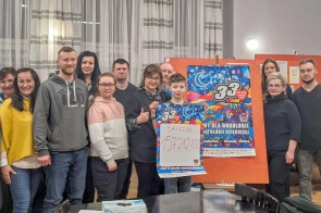 Grupa uśmiechniętych osób stoi razem, pozując do wspólnego zdjęcia w pomieszczeniu z jasnymi firanami w tle. W centralnej części chłopiec trzyma planszę z napisem „Skoczów 157.212,18”, obok kolorowy plakat finału WOŚP. Widać atmosferę zaangażowania i radości, podkreślającą sukces wspólnej inicjatywy na rzecz Wielkiej Orkiestry Świątecznej Pomocy.