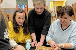 Trzy kobiety, trzecia od lewej, Beata Macura, Członkini Zarządu Powiatu, pochylają się nad stołem, na którym leży drewniana taca wypełniona monetami. Jedna z nich, ubrana w żółty sweter, ma na dłoni rękawiczkę ochronną. Kobieta liczy pieniądze. Towarzyszą jej dwie kobiety w czarnym i szarym ubraniu, uśmiechnięte i skupione na zadaniu. W tle widoczna jest puszka Wielkiej Orkiestry Świątecznej Pomocy oraz fragment sali.