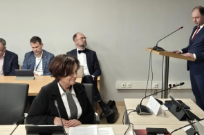 W sali konferencyjnej odbywa się XIX sesja Rady Powiatu. Adam Śliż, dyrektor Szpitala Śląskiego w Cieszynie, w granatowym garniturze, przemawia przy mównicy z mikrofonem, wyrażając swoje stanowisko. Janina Żagan, Starosta Cieszyński, w pierwszym planie, przegląda dokumenty z pełnym skupieniem. Na drugim planie kilku uczestników, elegancko ubranych, korzysta z tabletów. Atmosfera profesjonalna i poważna.