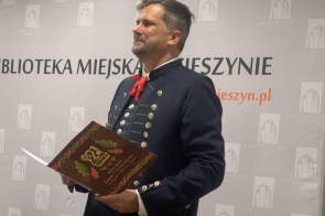 Dr Cezary Tomiczek, Prezes Macierzy Ziemi Cieszyńskiej w cieszyńskim ubiorze regionalnym, z charakterystycznymi zdobieniami i czerwonym krawatem, trzyma w rękach otwartą książkę z napisem „Macierz Ziemi Cieszyńskiej”. W tle widoczna jest ścianka promocyjna Biblioteki Miejskiej w Cieszynie, z logo instytucji i adresem strony internetowej „cieszyn.pl”. Mężczyzna patrzy w bok z lekkim uśmiechem.