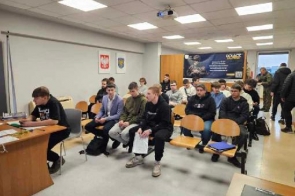 Grupa młodych mężczyzn siedzi w rzędach na drewnianych krzesłach w jasnej sali. W tle widoczne są symbole narodowe oraz powiatowe, a także duży baner promujący służbę wojskową. Po lewej stronie przy biurku pracuje urzędnik (nie jest widoczny na zdjęciu) z dokumentami. Pomieszczenie oświetlone jest jasnym światłem, a przez okno widać fragment zewnętrznego krajobrazu.
