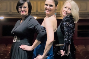 Trzy elegancko ubrane kobiety stoją na scenie teatru, ustawione w lekkim skosie, z dłońmi opartymi na biodrach. Ich postawy emanują pewnością siebie i pozytywną energią. Tło stanowi widownia z rzędem pustych, stylowych foteli oraz klasyczna, ozdobna architektura teatru. Oświetlenie nadaje scenie klimatyczny, ciepły nastrój, podkreślając wyjątkowy moment.