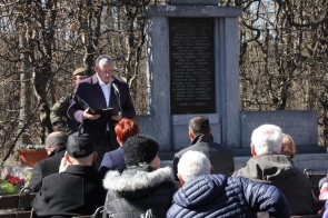 Adam Cieślar, dyrektor COK w Cieszynie w ciemnym płaszczu przemawia do mikrofonu przed pomnikiem z tablicą upamiętniającą poległych pod Wałką. W tle żołnierz w mundurze. Przed nim zgromadzeni ludzie w ciepłych kurtkach, siedzący na ławkach. Wokół gołe drzewa, ponieważ jest wczesna wiosna. 