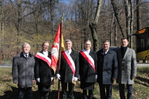 Sześć osób pozuje do zdjęcia w plenerze, na tle drzew i drogi – od lewej Stanisław Kubicius, Przewodniczący Rady, Katarzyna Stefka, Naczelniczka Wydziału Rady, Zarządu i Zdrowia, Krzysztof Glajcar, Kierownik Biura Transportu i Organizacji Ruchu, Maria Kowalska, Zastępczyni Naczelnika Wydziału Programowania i Funduszy Zewnętrznych, Jerzy Pilch, Członek Zarządu Powiatu oraz Jacek Wiśniewski, Sekretarz Powiatu. Trzy osoby w czarnych płaszczach i białych rękawiczkach trzymają sztandar z orłem. Towarzyszą im trzej mężczyźni w eleganckich płaszczach. W tle żółty autobus z napisem „UROCZYSTY PRZEJAZD POD WAŁKĘ”.