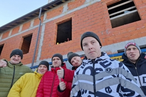 Grupa sześciu mężczyzn w zimowych kurtkach i czapkach pozuje na tle budowy z czerwonej cegły. Niektórzy uśmiechają się i pokazują kciuki w górę. W tle widać dźwig budowlany i otwarte okna w niedokończonym budynku. Atmosfera radosna i pełna energii.