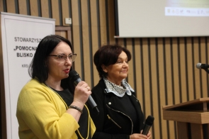 Dwie kobiety z mikrofonami przemawiają podczas Konferencji „Żyj realnie, nie wirtualnie”. Jedna w żółtym swetrze (Ilona Fajter-Kruczek, Prodziekan ds. Kształcenia UŚ w Cieszynie), druga w czarnej marynarce z apaszką (Janina Żagan, Starosta Cieszyński). W tle drewniana ścianka i baner Stowarzyszenia „Pomocna Dłoń – Blisko Ciebie”.
