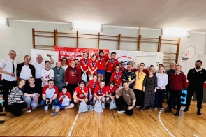 Grupa uczestników i organizatorów XV Beskidzkiego Mityngu w Trójboju Siłowym pozuje wspólnie w sali gimnastycznej. W centrum zawodnicy w czerwonych koszulkach z medalami, w tle banery Olimpiad Specjalnych i partnerów wydarzenia.