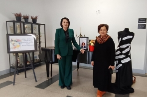 Dwie kobiety (od lewej Kornelia Zubek, dyrektor Zespołu Szkół Gastronomiczno-Hotelarskich im. W. Reymonta w Wiśle, od prawej Janina Żagan, Starosta Cieszyński) stoją przy fortepianie w szkolnym holu; obok nich czarna suknia z hasłami opisującymi kobiecą siłę i tablica z napisem „Ona – kobieta, bohaterka, autorka, sztuka, muza. Rzecz o herstorii”. W tle gablota z pucharami.