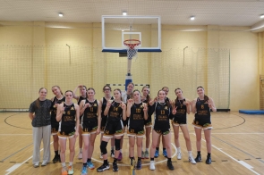 Drużyna koszykarek Liceum Ogólnokształcącego im. Pawła Stalmacha w Wiśle świętuje sukces na hali sportowej. Zawodniczki ubrane w czarne stroje z napisem „Stalmach Basketball” prezentują medale i wykonują gesty zwycięstwa, uśmiechając się do zdjęcia.