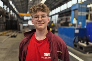  Mateusz Sikora, ubrany w czerwoną koszulkę i uniform szkoły technicznej, z uśmiechem stoi w przestronnej hali fabrycznej. W tle widoczne są maszyny i konstrukcje przemysłowe. Jest zadowolony z warunków w trzynieckim zakładzie.