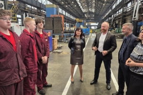 Grupa osób w hali fabrycznej rozmawia i uśmiecha się. Po lewej trzech uczniów w czerwonych uniformach, po prawej dyrektor Bożena Cholewa, prezes Marian Czyż i dwie inne osoby w eleganckich strojach, stoją w luźnej rozmowie.
