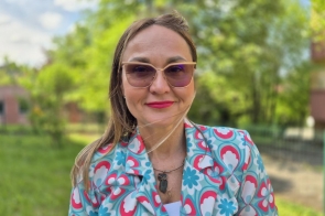 Dorota Prysak, pracownik Wydziału Sztuki i Nauk o Edukacji, na zdjęciu prezentuje się w kolorowym kostiumie o wzorach w odcieniach różu i błękitu. Ma na sobie okulary przeciwsłoneczne i uśmiech na twarzy. Tło stanowi zielony krajobraz z drzewami, co dodaje radosnej atmosfery.