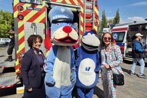 Od lewej strony znajdują się Janina Żagan, starosta cieszyński, oraz Dorota Prysak, obie uśmiechnięte. Obok nich stoi osiołek w kostiumie maskotki, co nadaje radosny klimat całej scenie. W tle widoczne są pojazdy ratunkowe, co wskazuje na wydarzenie o tematyce bezpieczeństwa.