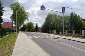 Droga z pasami dla pieszych prowadzi wzdłuż ogrodzonych posesji i drzew. Po lewej stronie jest chodnik, a w tle czerwony dach domu. Nad przejściem dla pieszych znajdują się znaki i lampy. Na niebie widać chmury i niebieskie niebo.