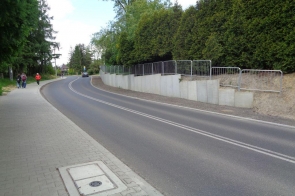 Asfaltowa droga z podwójną linią biegnie wzdłuż betonowego muru i żywopłotu. Po lewej stronie widoczny jest chodnik, na którym idą ludzie. W tle droga zakręca, a na niej widać samochód i znaki drogowe