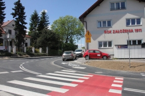 Przejście dla pieszych z czerwonym pasem, prowadzące do zakrętu. Po prawej budynek z napisem „ROK ZAŁOŻENIA 1911”. W tle stoją samochody i drzewa. Znaki drogowe wskazują na obecność rowerzystów. Okolica jest spokojna i zadbana.