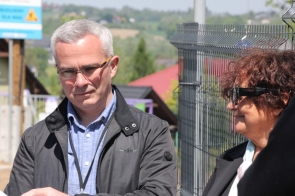 Od lewej - Przemysław Kulawiec, Kierownik Zespołu Inwestycji PZDP w Cieszynie i Janina Żagan, Starosta Cieszyński stoją obok siebie na tle ogrodzenia. Mężczyzna ma siwe włosy i okulary, kobieta ciemne okulary przeciwsłoneczne. W tle widoczny jest rozmyty krajobraz z domami i wzgórzami. Atmosfera jest spokojna i słoneczna.