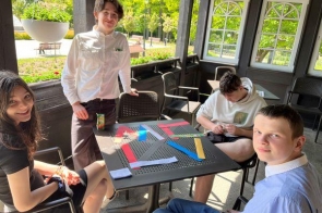 Czworo młodych osób siedzi przy stole na werandzie w słoneczny dzień. Uśmiechają się i pracują z kolorowymi paskami papieru. W tle widać drzewa i park. Atmosfera jest swobodna i twórcza.