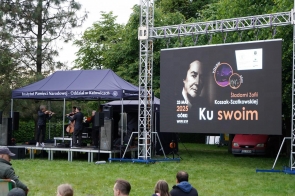 Pod sceną ustawioną na trawie kilka osób słucha występu kwartetu smyczkowego grającego pod namiotem Instytutu Pamięci Narodowej. Obok widnieje duży ekran z grafiką wydarzenia „Ku swoim. Śladami Zofii Kossak-Szatkowskiej”.