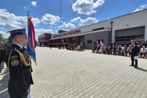 Uroczystość odbywa się przed budynkiem Komendy Powiatowej Państwowej Straży Pożarnej. Strażacy w galowych mundurach stoją w szeregu, po przeciwnej stronie siedzą zaproszeni goście, a centralnie widoczny jest sztandar i prowadzący wydarzenie.