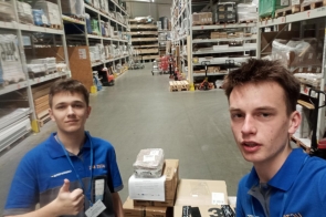 Franek Olszar i Mateusz Moskała, ubrani w niebiesko-szare koszulki z napisem „UCZEŃ”, stoją w przestronnej hali magazynowej między wysokimi regałami. Franek robi selfie, a Mateusz pokazuje gest uniesionego kciuka. Przed nimi ułożone są kartony.
