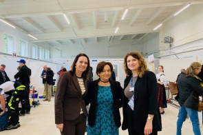 Trzy kobiety - w środku Janina Żagan, Starosta Cieszyński, po lewej - Sylwia Cieślar, Wójt Goleszowa stoją razem w jasnej sali z białymi boksami konkursowymi w tle. Ubrane elegancko, uśmiechają się do aparatu. W tle widoczni uczestnicy wydarzenia, osoby pracujące przy stanowiskach i goście.