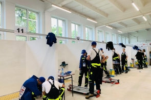 Uczestnicy ubrani w robocze stroje i niebieskie czapki pracują w boksach oznaczonych numerami, skupiając się na zadaniach technicznych. Widać narzędzia, sprzęt i stanowiska pracy, co wskazuje na zawody praktyczne w hali szkoleniowej.