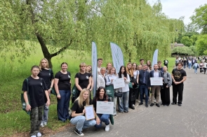 Grupa młodzieży i nauczycieli pozuje na tle zielonych drzew z dyplomami i tablicami informującymi o nagrodzie w konkursie „Zielona Pracownia”. Uczestnicy są uśmiechnięci, w tle widoczni są spacerujący ludzie i drewniana chata.
