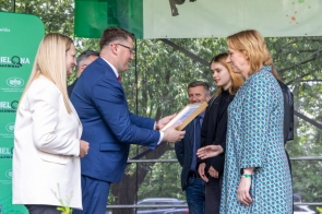 Mężczyzna w garniturze wręcza dyplom kobiecie w kolorowej sukience; obok niej stoi młoda dziewczyna. W tle uśmiechnięci uczestnicy wydarzenia i zielona ścianka promująca projekt „Zielona Pracownia”.