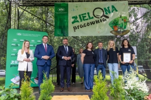Grupa osób stoi na scenie pod zielonym banerem „Zielona pracownia”. Uśmiechnięci przedstawiciele szkół i instytucji, niektórzy w garniturach, inni w koszulkach z logo projektu, jedna z kobiet trzyma dyplom. W tle zieleń drzew i dekoracje roślinne.