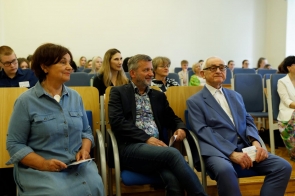 Osoby siedzące w pierwszym rzędzie sali audytoryjnej, wśród nich starszy mężczyzna w niebieskim garniturze, Michał Rajwa, Naczelnik Wydziału Edukacji w marynarce i Janina Żagan, Starosta w niebieskiej sukience. W tle publiczność, głównie młodzież szkolna.