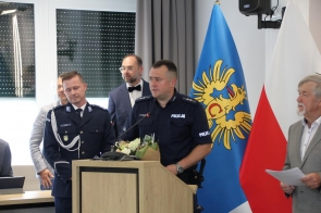 Policjant w granatowym mundurze przemawia przy mównicy, trzymając bukiet kwiatów. Obok stoją inni funkcjonariusze oraz przedstawiciele władz. W tle widoczne są flagi Powiatu Cieszyńskiego i Polski. Uroczysta, oficjalna atmosfera.
