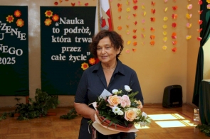Starosta Janina Żagan uśmiecha się, trzymając bukiet różowych i białych kwiatów. Za nią dekoracja z cytatem o nauce i kolorowymi motylami na ścianie, w tle sztandar oraz plansze zakończenia roku.