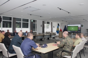 Sala konferencyjna Starostwa Powiatowego w Cieszynie. Przy długich stołach siedzą przedstawiciele służb, wojska, policji i samorządu. W tle ekrany z flagami i transmisją online oraz okna z zasłonami.