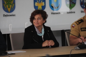 Janina Żagan, Starosta Cieszyński, siedzi przy stole konferencyjnym, uśmiecha się lekko i spogląda w bok. Za nią widoczne są herby gmin powiatu: Dębowiec, Hażlach, Istebna, Skoczów.