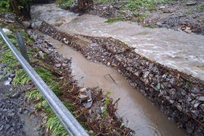 Uszkodzona konstrukcja z koszy siatkowo-kamiennych przy drodze powiatowej. Wysoki poziom wody w potoku podmywa umocnienia, a nurt niesie rumosz. Widać liczne zniszczenia pobocza i wypłukaną ziemię.