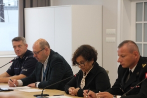Janina Żagan, Starosta Cieszyński, prowadzi obrady w otoczeniu przedstawicieli służb: policjanta, urzędnika i brygadiera Michała Pokrzywy z PSP. Wszyscy notują i mówią do mikrofonów. W tle jasne wnętrze z oknem zaklejonym taśmą anonimizującą.