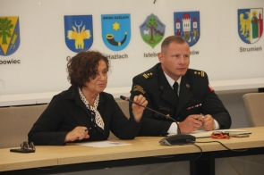 Janina Żagan, Starosta Cieszyński, mówi do mikrofonu podczas posiedzenia, trzymając długopis. Obok niej siedzi brygadier Michał Pokrzywa w mundurze PSP. Za nimi widoczne herby gmin powiatu cieszyńskiego.