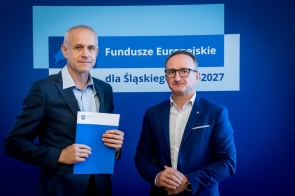 Jacek Michałek, naczelnik Wydziału Programowania i Funduszy Zewnętrznych Starostwa Powiatowego w Cieszynie, w eleganckim garniturze trzyma niebieską teczkę z dokumentami. Obok niego stoi Grzegorz Boski, wicemarszałek Województwa Śląskiego. Oboje pozują na tle tablicy „Fundusze Europejskie dla Śląskiego 2021–2027”, podkreślając formalny charakter wydarzenia.