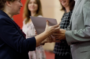 Osoba wręcza dokument drugiej osobie, w tle stoją dwie uśmiechnięte kobiety.