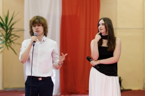 Dwójka młodych osób z mikrofonami stoi na tle biało-czerwonej dekoracji.