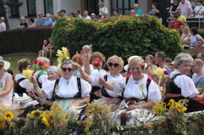 Grupa kobiet w tradycyjnych strojach ludowych siedzi na udekorowanym wozie z kwiatami i kłosami zbóż podczas Dożynek Powiatowo-Gminnych w Goleszowie.