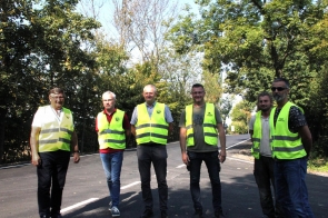 Sześciu mężczyzn stoi na świeżo wyremontowanej drodze otoczonej zielonymi drzewami. Większość ubrana jest w odblaskowe, żółte kamizelki, a część ma na sobie koszule lub koszulki.