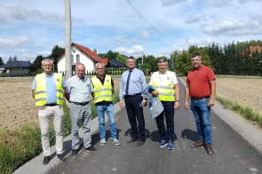 Sześciu mężczyzn stoi na świeżo wyremontowanej, asfaltowej drodze. Część z nich ubrana jest w odblaskowe kamizelki, inni mają na sobie koszule lub koszulki. Obok asfaltu widać pole uprawne i rząd drzew, a w tle znajdują się domy jednorodzinne z czerwonymi dachami. Pogoda jest słoneczna z lekkim zachmurzeniem.