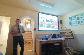 Osoba w mundurze stoi obok dużego, jasnego ekranu z kolażem zdjęć. Po lewej łukowe przejście ze schodami. Przed ścianą biurko z laptopem i sprzętem; na ścianach obrazy, na suficie listwa oświetleniowa.