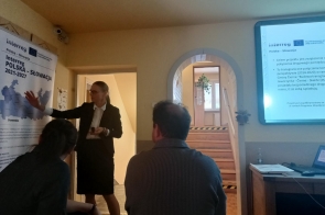 Kobieta przy tablicy wskazuje element; obok siedzą dwie osoby tyłem. W tle łukowe przejście ze schodami i rośliną. Po prawej krawędź jasnego ekranu prezentacji; na ścianie półki i materiały.